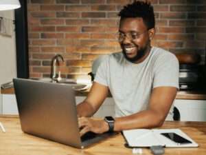 Benjamin Farin, Social Media Experte, sitz in der Küche am Tisch und am Arbeitet an seinem silbernem Laptop und ist am Lachen. Trägt Brille und graues Hemd.